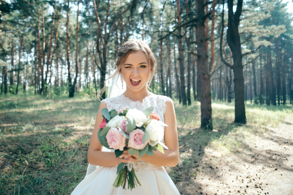 Winking bride in wedding dress holding bouquet on natural background
